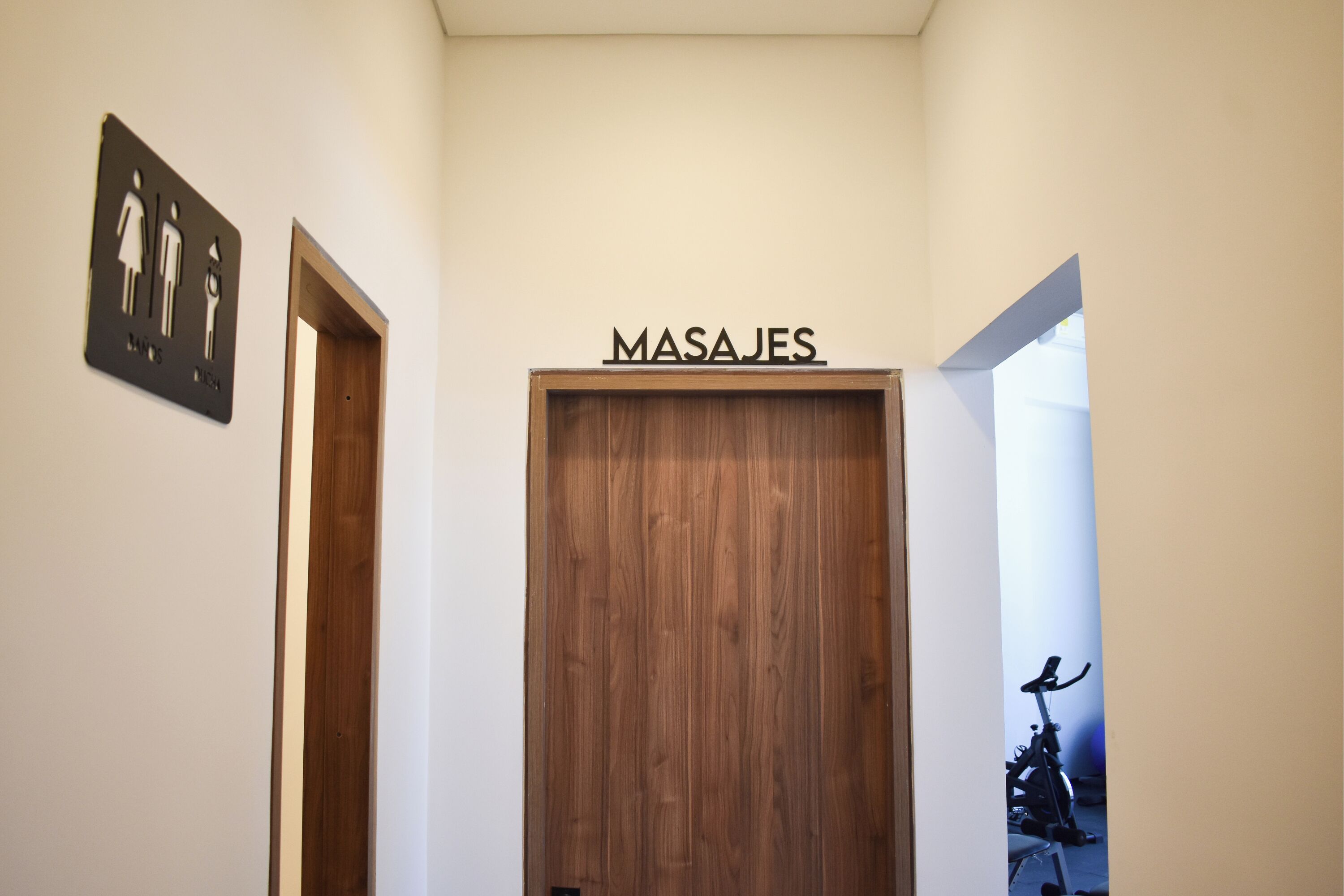 Un couloir avec une porte en bois portant l’inscription « MASAJES » et un panneau indiquant les toilettes pour hommes et pour femmes.