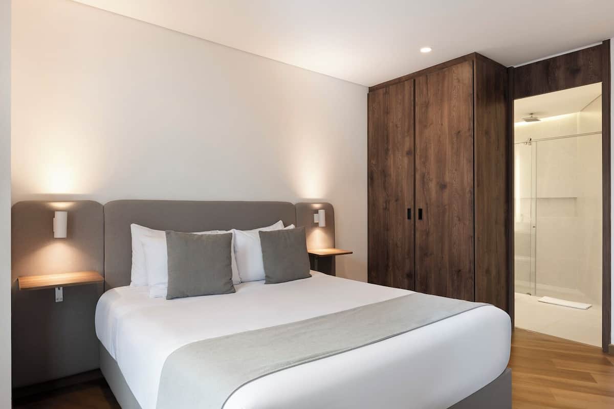 Una habitación de hotel moderna con una cama, mesitas de noche, luces fijadas a la pared, un armario y un box de ducha de cristal.