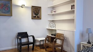 Una habitación con vigas de madera, paredes blancas, una estantería, un escritorio, dos sillas y una cama con cubrecama estampado.