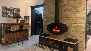 Una sala de estar moderna con pared de piedra, chimenea negra, zona de bar de madera y suelo de baldosas.