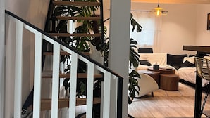 Una escalera de madera con barandas blancas conduce a una sala de estar con un sofá, una mesita de centro y plantas en macetas.