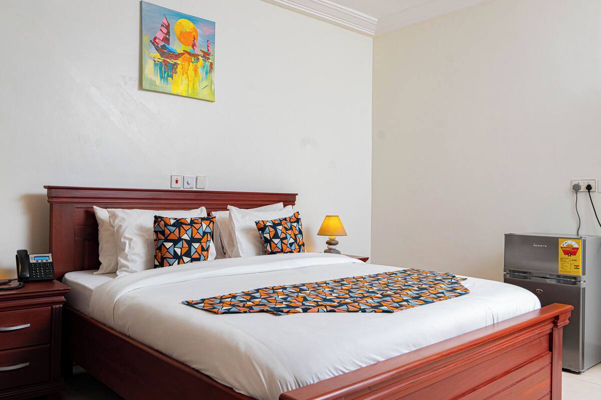 Cama bem arrumada com lençóis brancos e almofadas estampadas, cabeceira de madeira, mesa de cabeceira com abajur, telefone e um pequeno aquecedor.