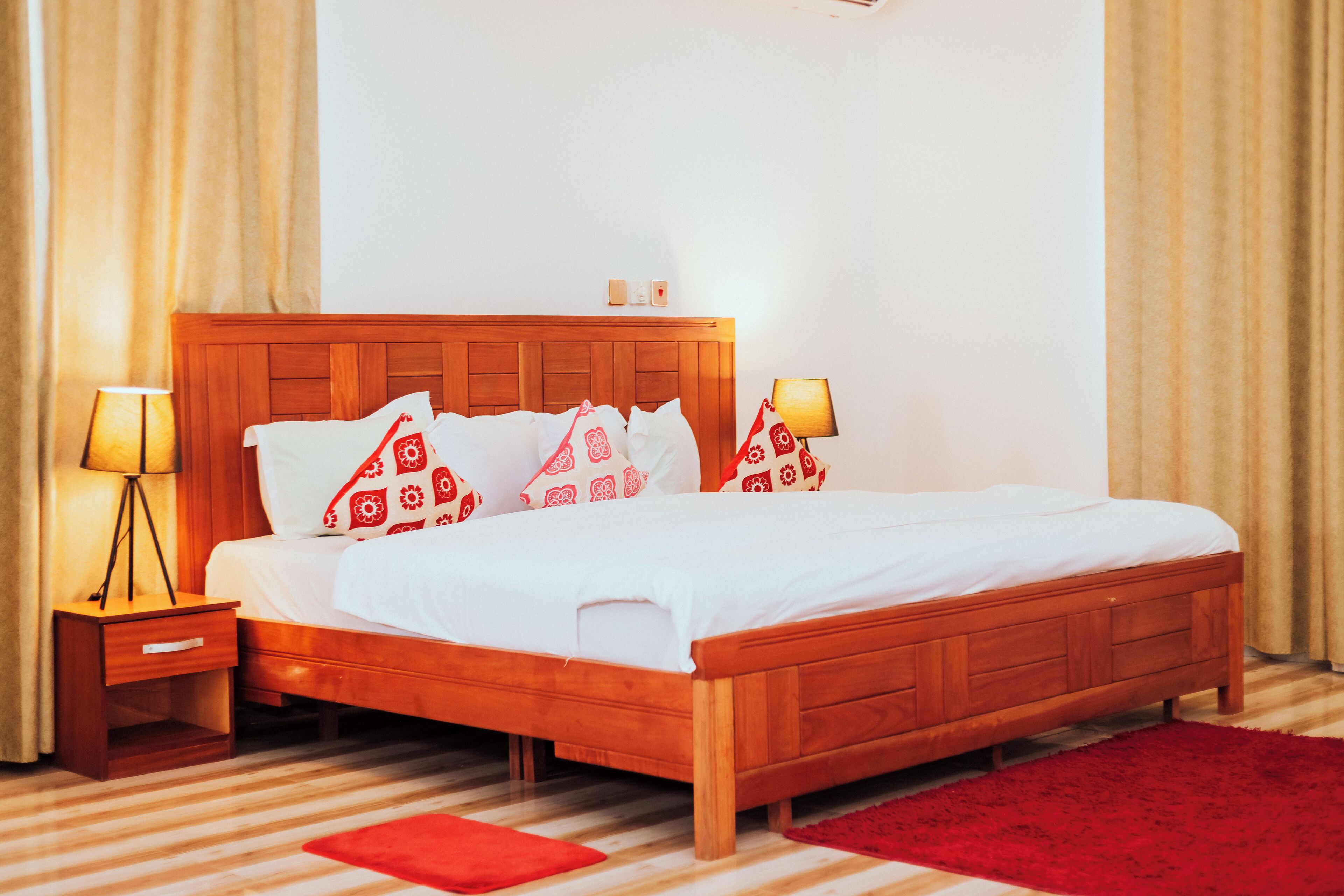 A wooden bed with white bedding and red patterned pillows, a bedside table with a lamp, and a red rug on a striped floor.