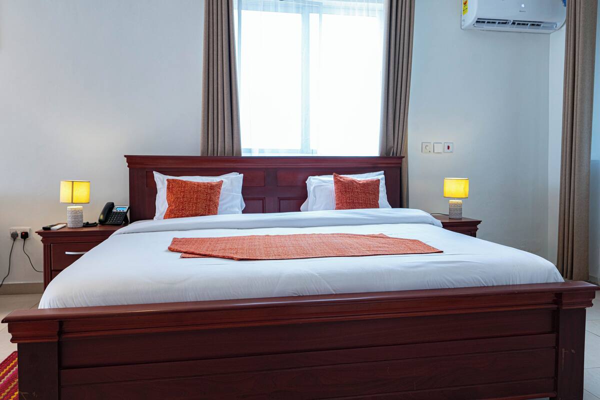 Quarto de hotel com cama de madeira, roupa de cama branca, duas luminárias de mesa, telefone e unidade de ar condicionado.