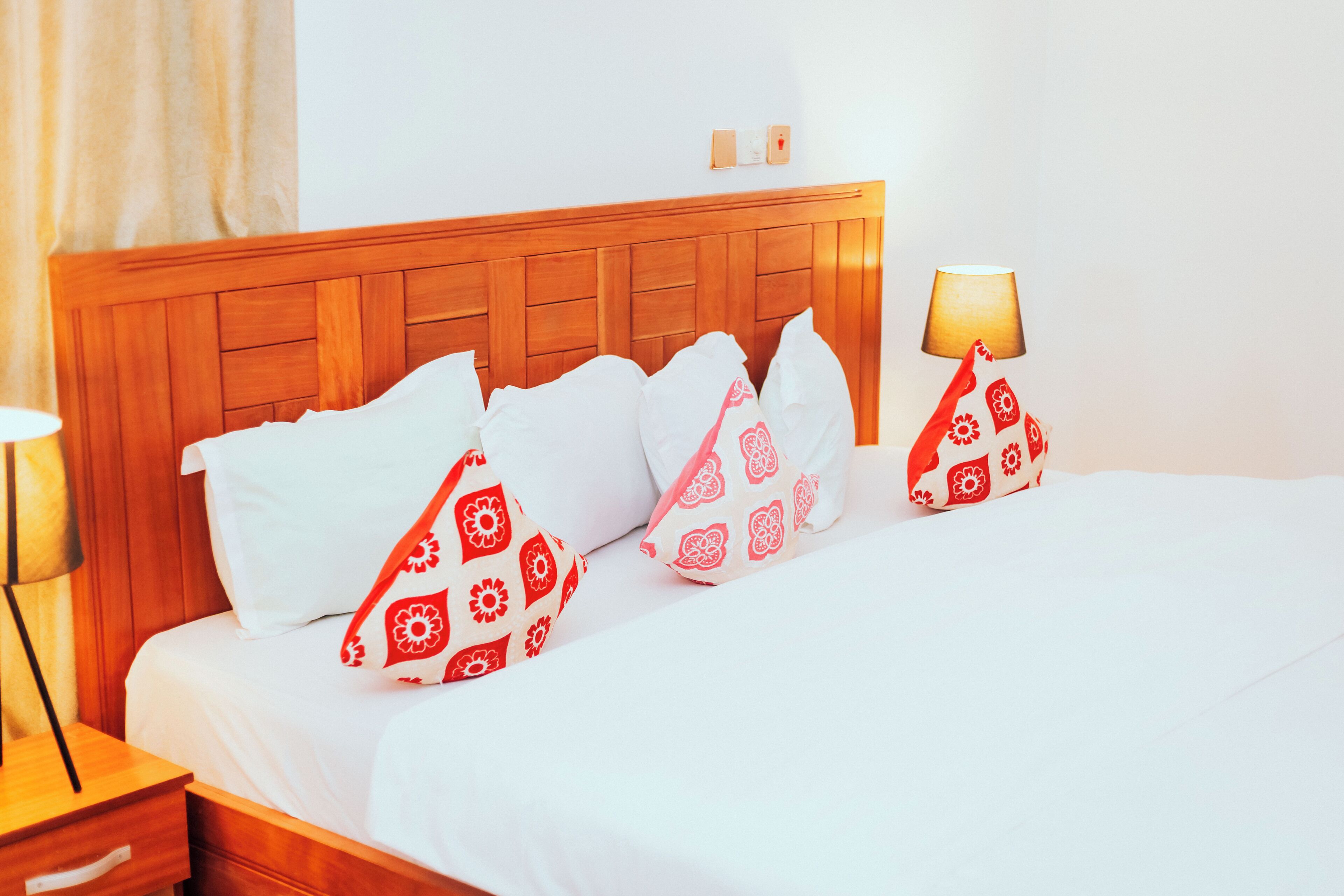 Una cama bien hecha con ropa de cama blanca y almohadas estampadas en rojo, un cabecero de madera, una lámpara de noche y una mesita de noche con un cajón.