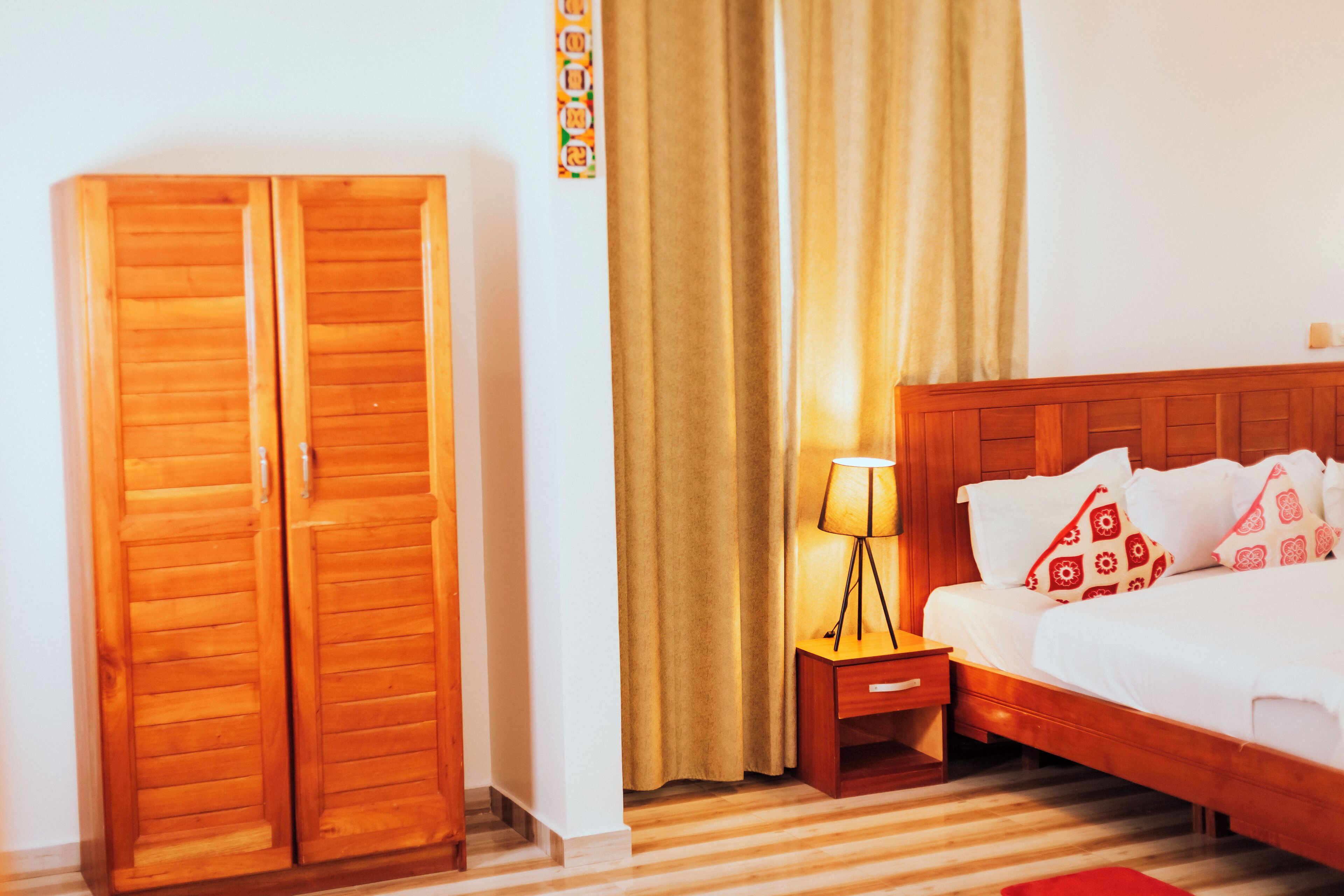 A hotel room with a wooden wardrobe, a bed with white bedding and red patterned pillows, a nightstand with a lamp, and a striped carpet.