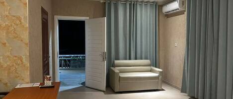 A room with a wooden desk, a beige sofa, a ceiling fan, and a view through a door to an outdoor area.