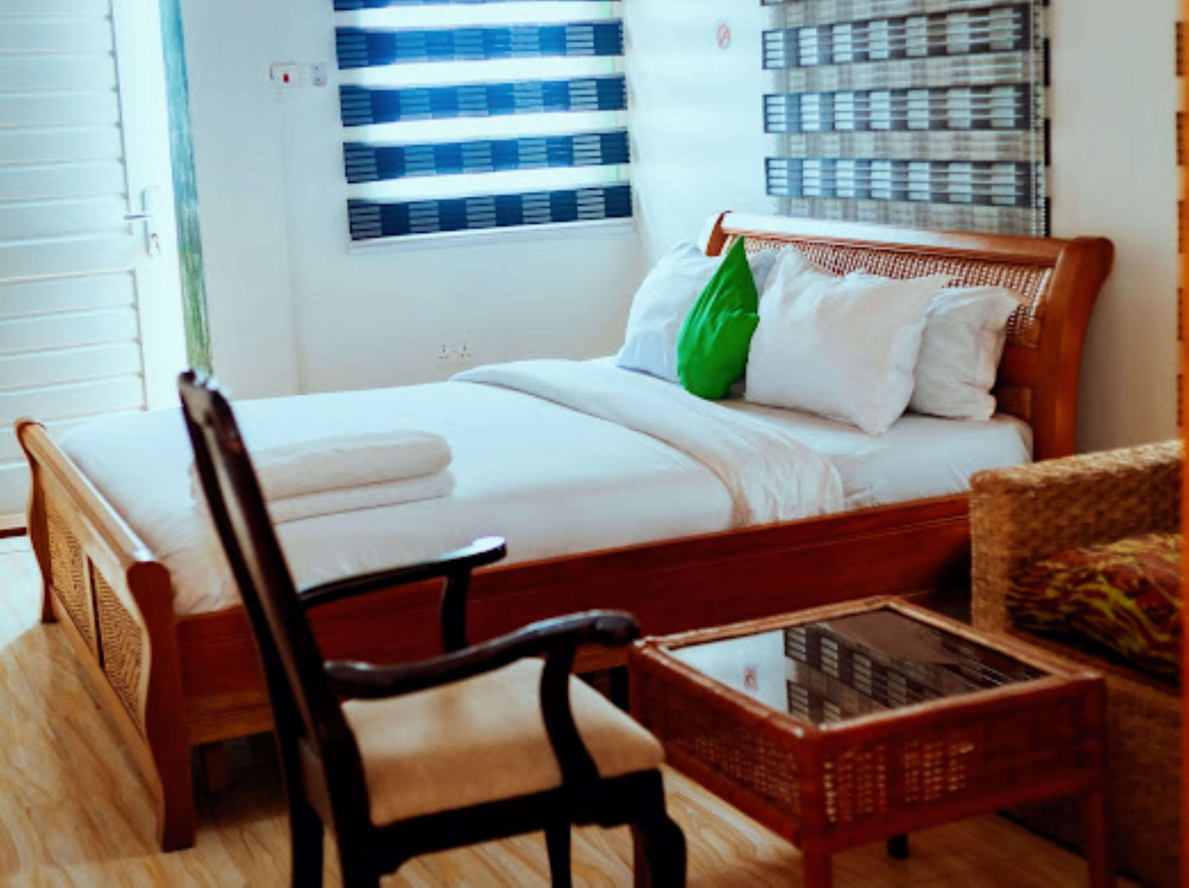 Habitación de hotel ordenada con una cama de madera, una silla, una mesita y una ventana con persianas.