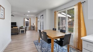 A modern living room with a wooden dining table, black chairs, a sofa, and a TV. There is a large window with curtains, a door leading to another room, and a small table with a plant.