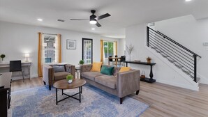 A modern living room with a grey sofa, a coffee table, and a ceiling fan. There is a staircase with a black railing leading to an upper level.