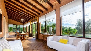Una habitación amplia con techo alto, ventanas grandes, un sofá blanco con un cojín amarillo y un comedor con mobiliario de madera.