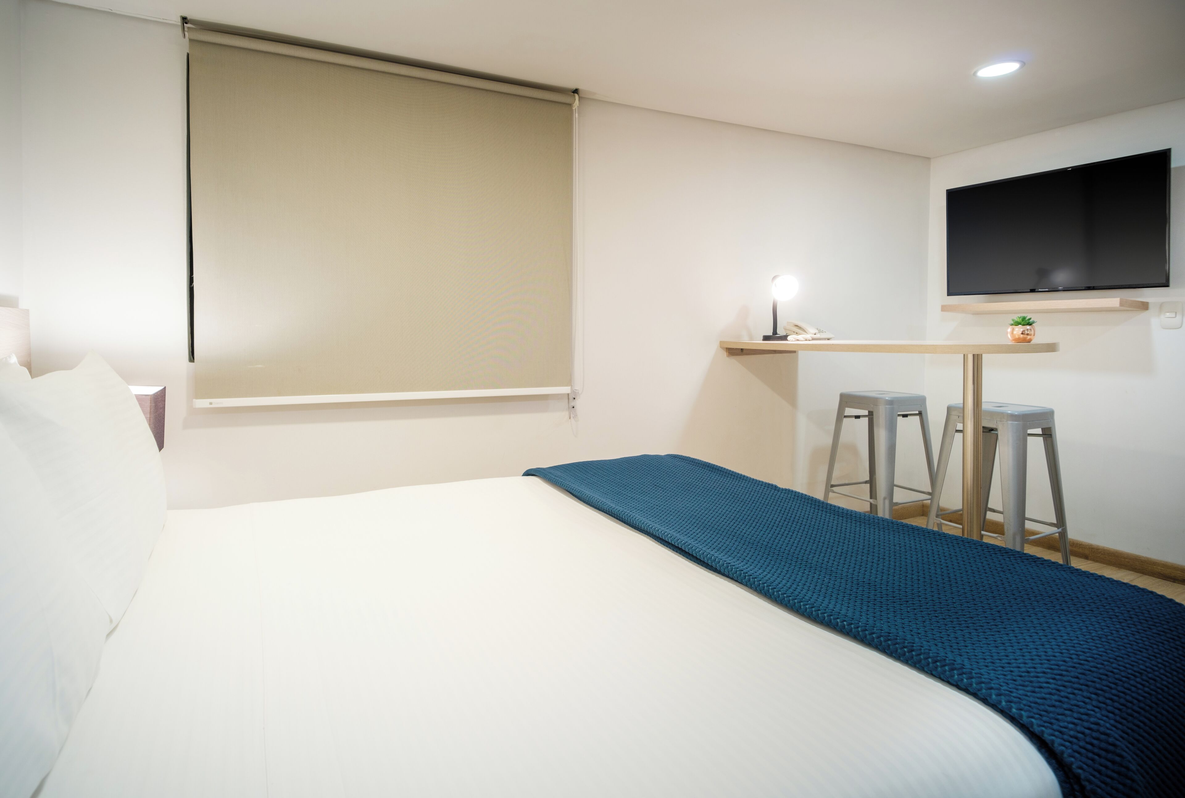 Habitación de hotel con cama, escritorio con dos taburetes, televisor montado en la pared y ventana con persiana.