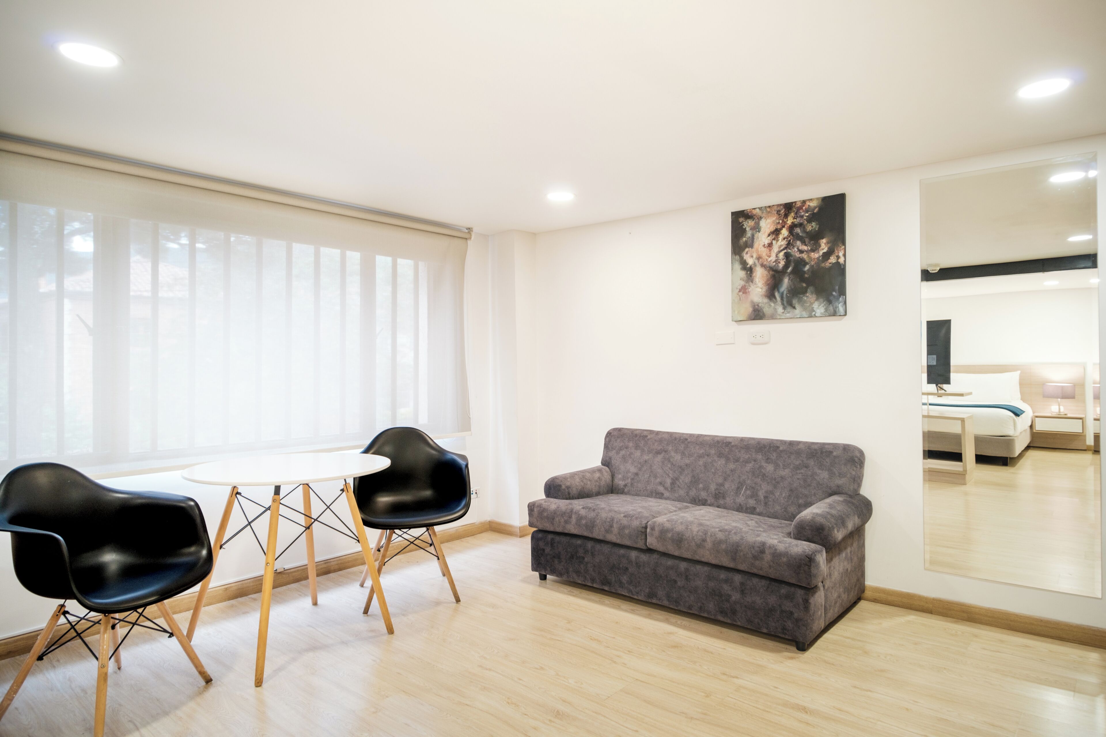 A modern room with a grey sofa, two black chairs, a round table, and a large window with blinds.