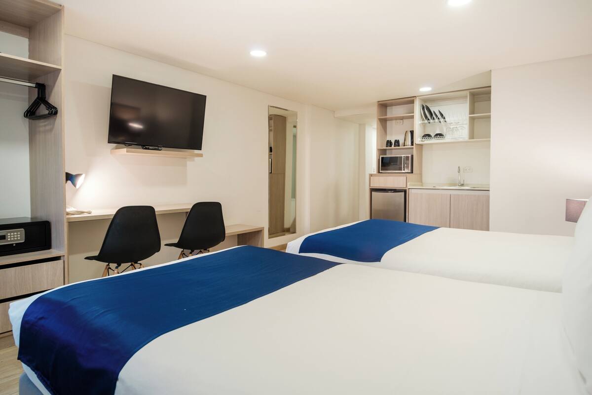 Ett hotellrum med två sängar, ett skrivbord, en TV och ett litet kök.