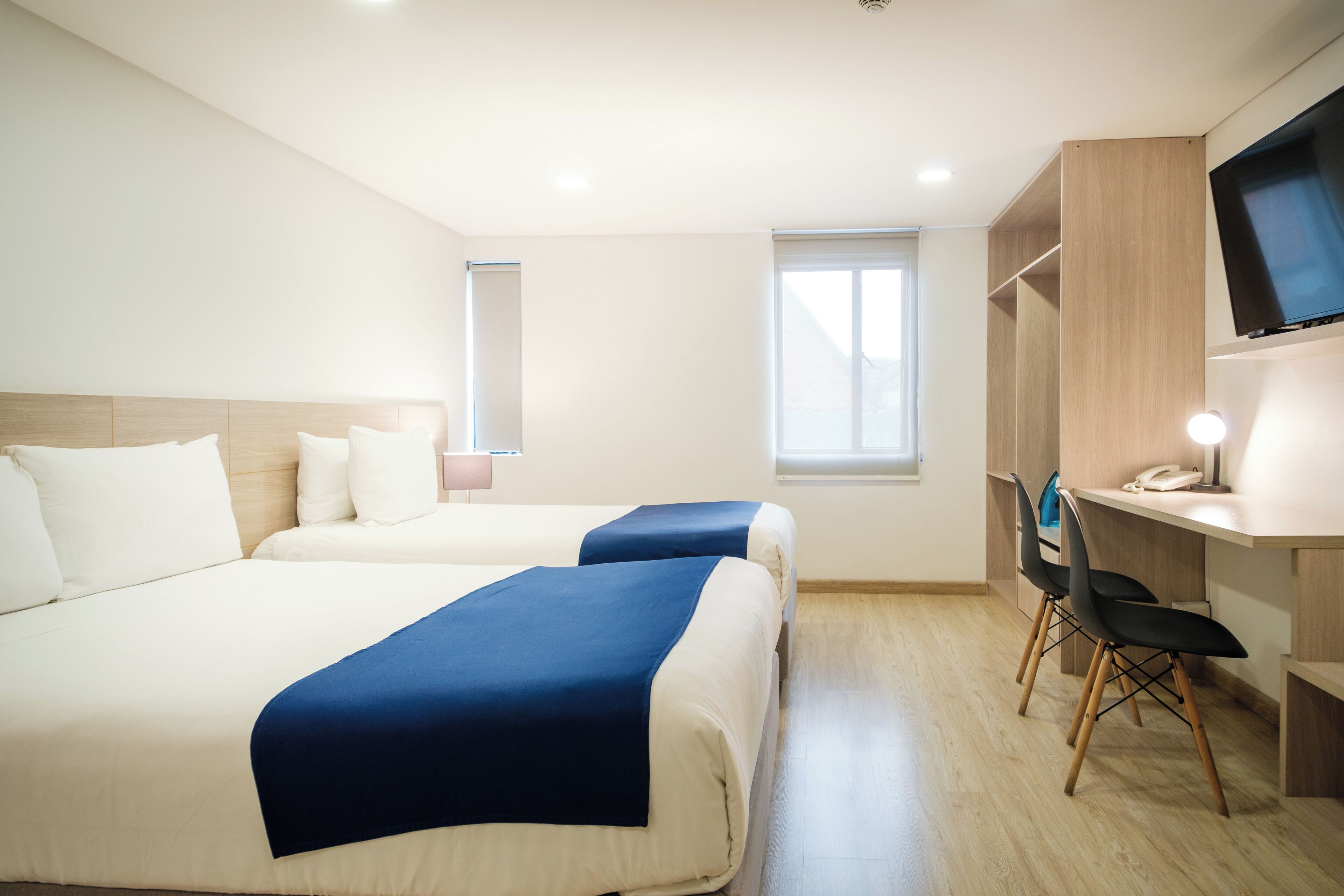 Habitación de hotel con dos camas, un escritorio con silla, televisor montado en la pared y ventana que proporciona luz natural.