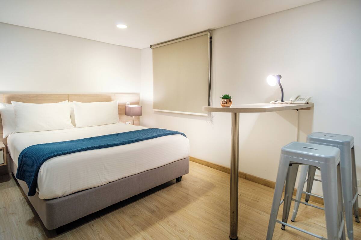 Ett hotellrum med en säng, ett nattduksbord, en lampa och ett litet skrivbord med en växt och en telefon.
