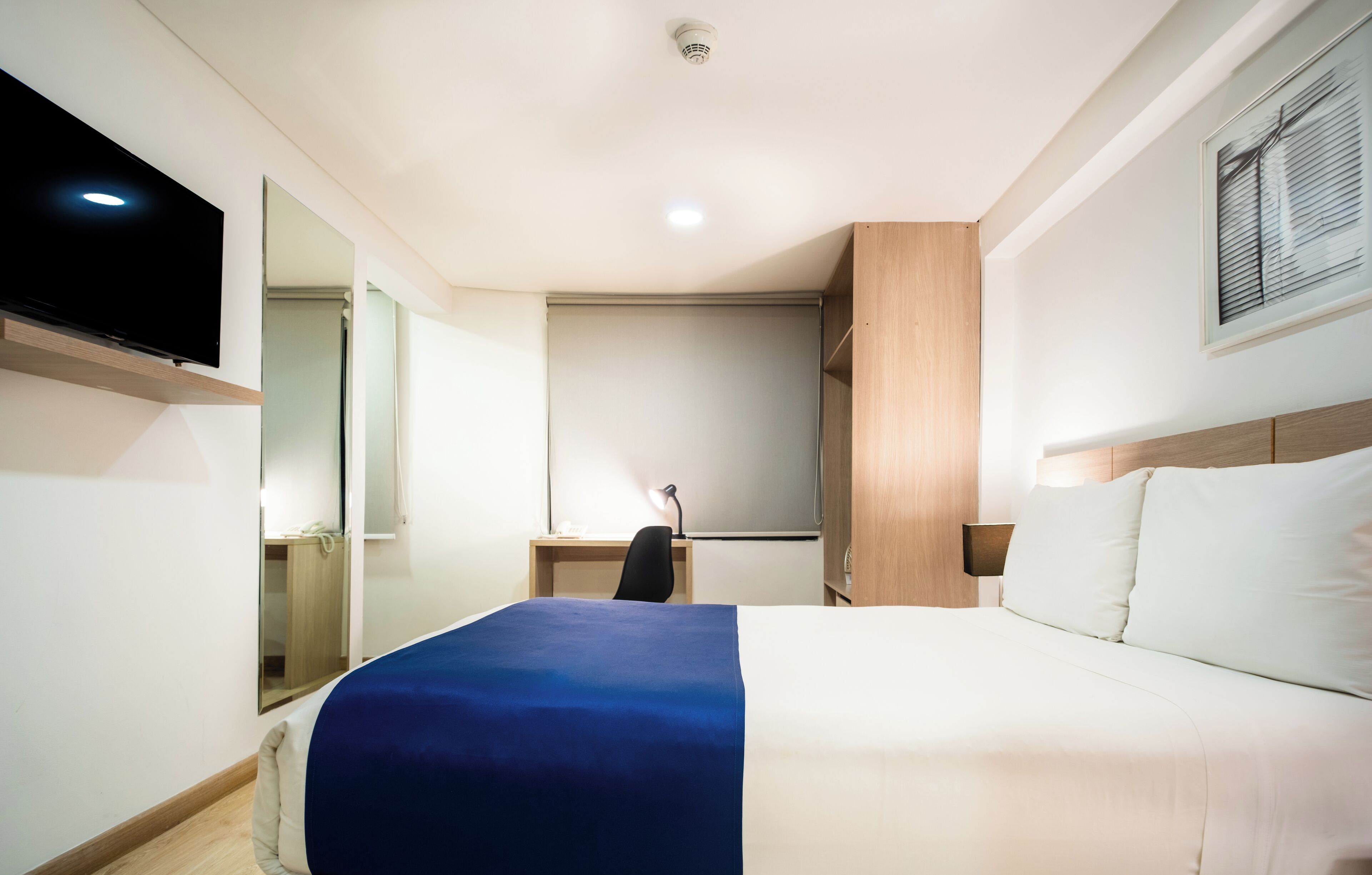 A hotel room with a bed, a desk, a chair, a TV, and a mirror.