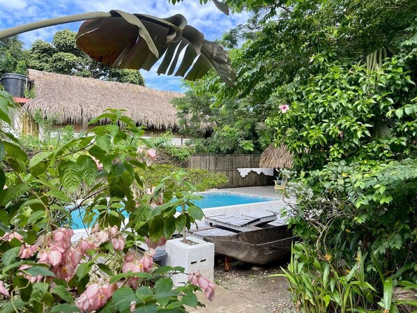 A backyard with a swimming pool, a thatched-roof structure, and lush greenery.