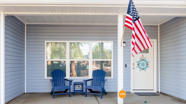Eine Veranda mit zwei blauen Adirondack-Stühlen, einem kleinen Tisch und einer amerikanischen Flagge.