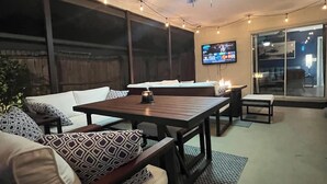 Un espace de vie extérieur comprenant un canapé, une table et des chaises, un ventilateur de plafond, des guirlandes lumineuses et un téléviseur fixé au mur.