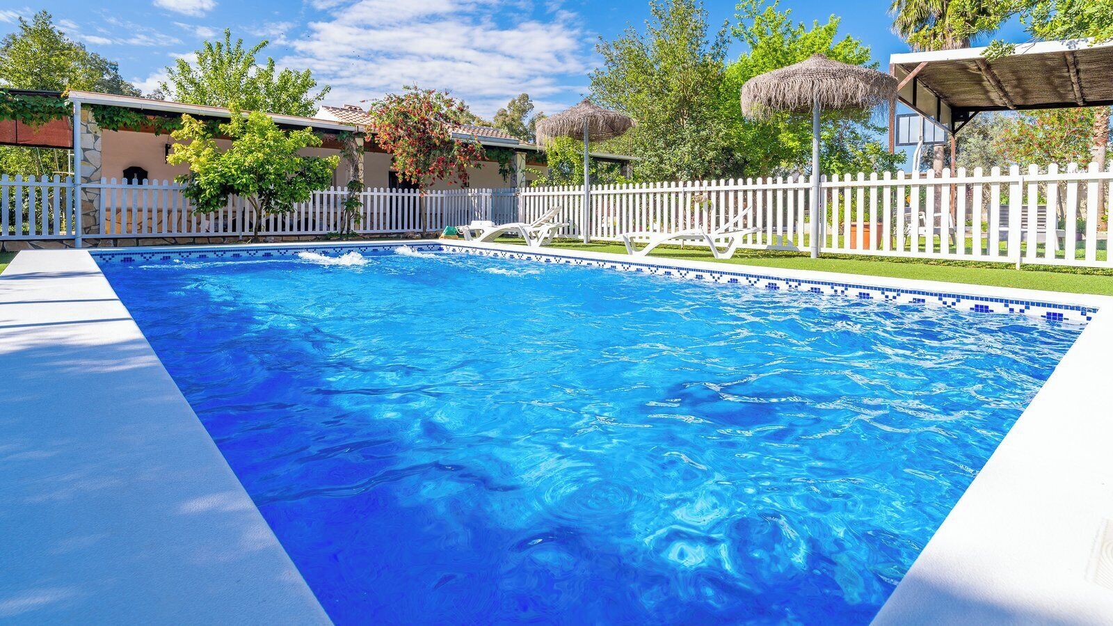 A swimming pool with a white fence, lounge chairs, and a thatched-roof structure.