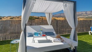 Una cama con dosel blanco, con una mesita, bebidas y un sombrero, frente a un seto de madera y un paisaje montañoso.