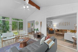 Uma sala de estar espaçosa com um sofá cinzento, mesa de centro de madeira e cadeiras brancas. Há uma janela grande que oferece vista para o exterior.
