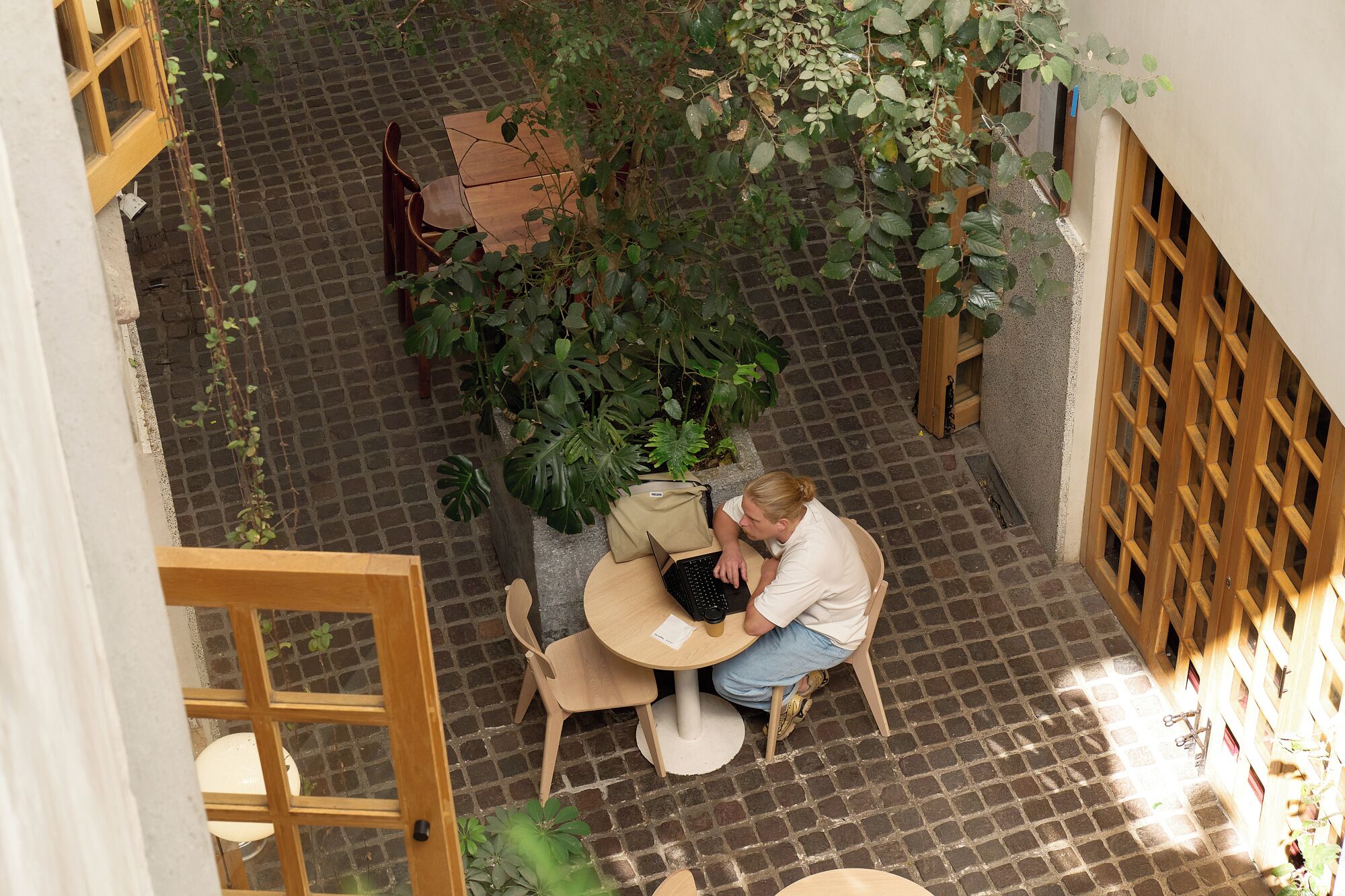A person seated at a round table with a laptop, surrounded by greenery and a brick floor.