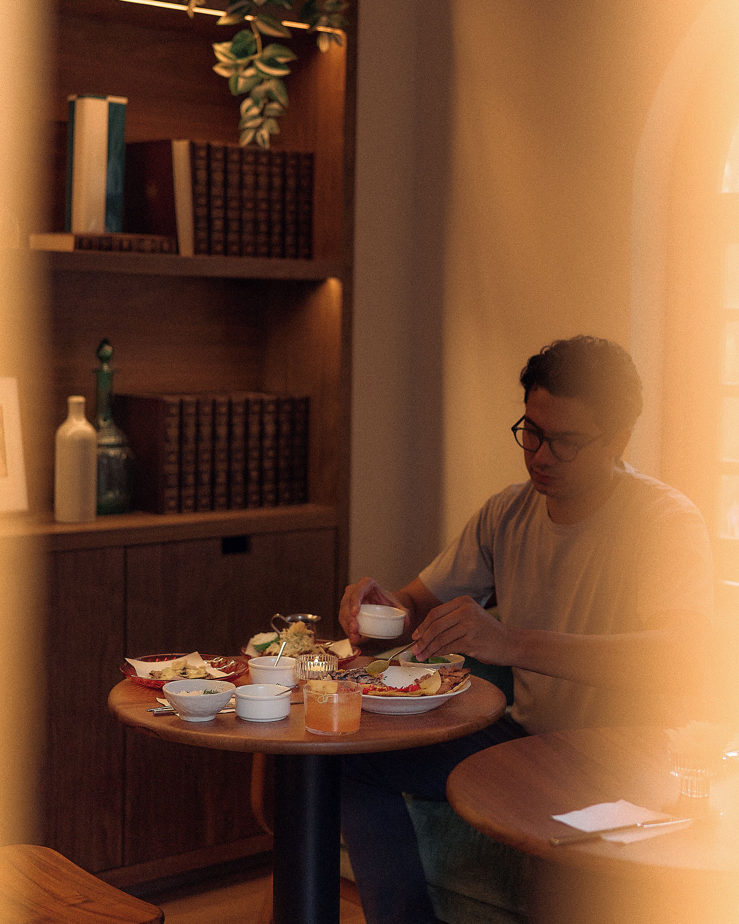Una persona sentada a una mesa redonda con varios platos, una bebida y una estantería al fondo.