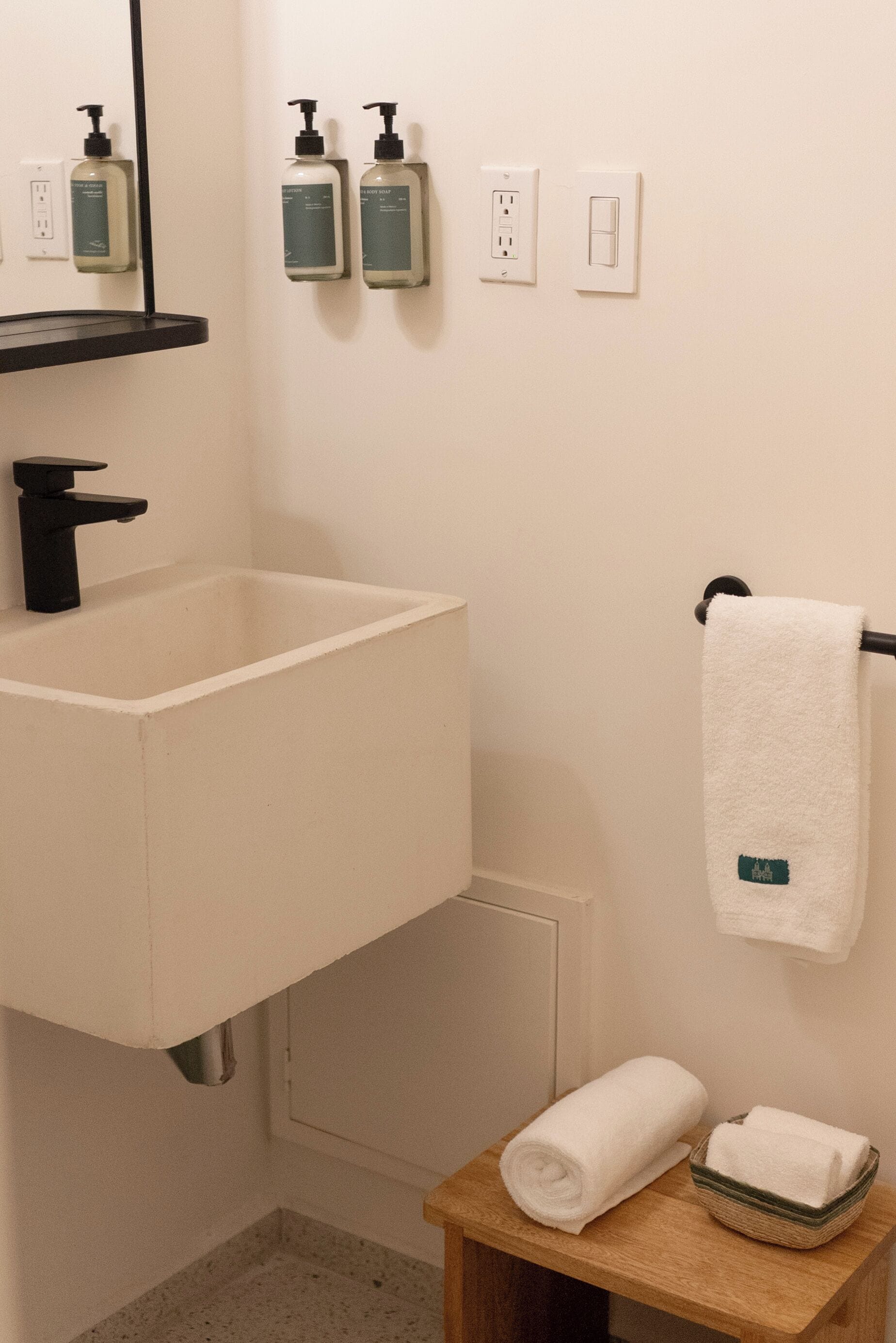 Une salle de bain équipée d’un lavabo, de deux distributeurs de savon, d’un porte-serviettes et d’une petite table en bois sur laquelle est posée une serviette.