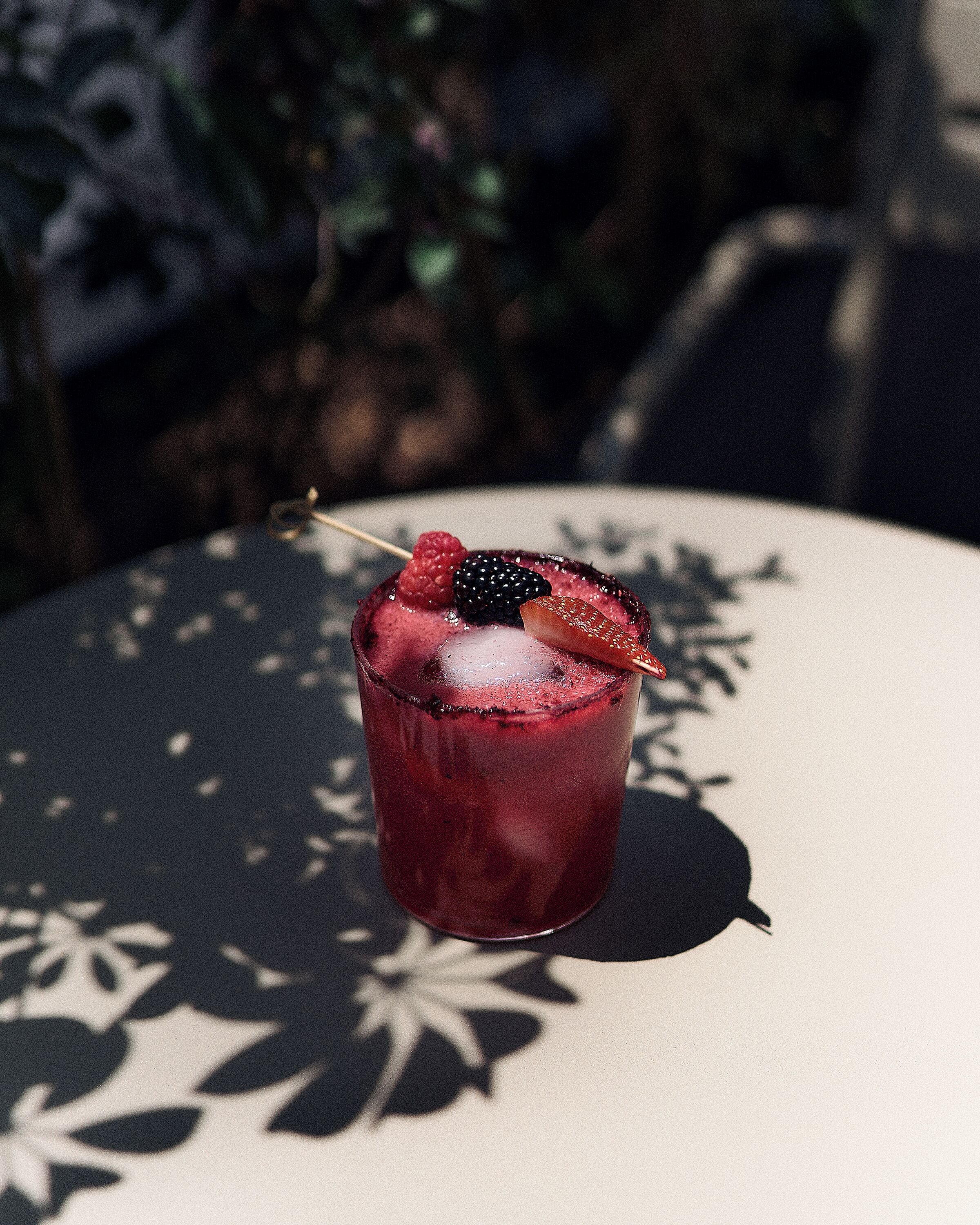 A glass of red beverage with berries and a straw on a table with a shadow pattern.