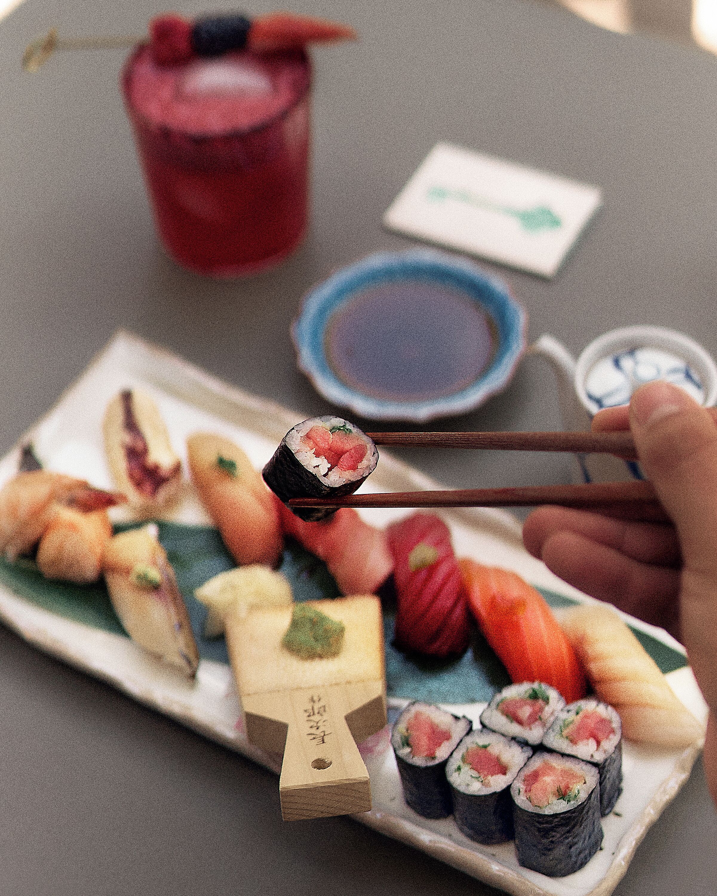 A plate of sushi with chopsticks picking up a piece, a red drink, and a small bowl of soy sauce on a table.