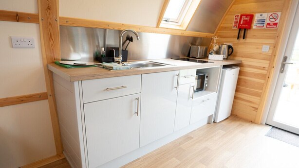 A compact kitchen with white cabinets, a stainless steel sink, and a small refrigerator. - Cosy Spacious Cabin with Wood Fired Hot Tub (Cumwhitton)