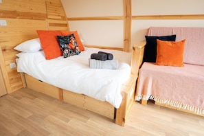 A wooden bed with white bedding and orange pillows, a small wooden nightstand, and a pink sofa with a blanket.