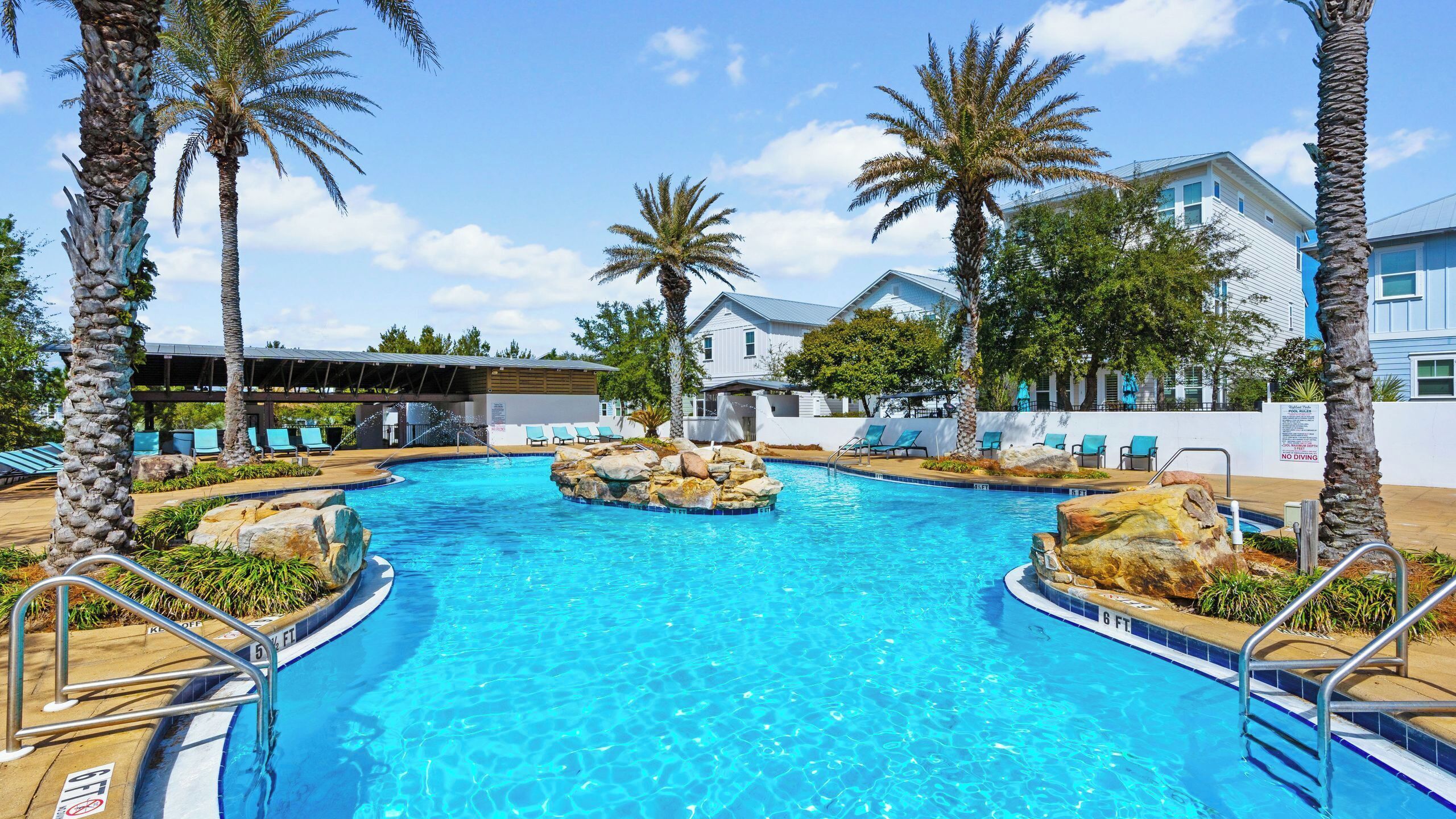 A clear blue pool with palm trees, lounge chairs, and a building in the background.