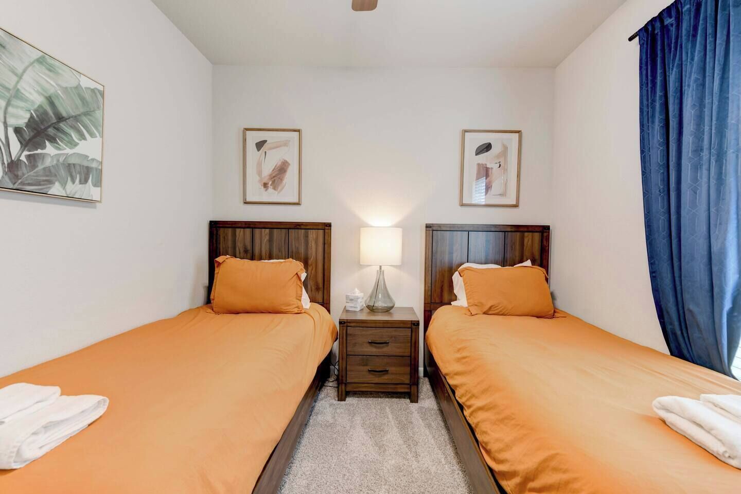 A hotel room with two single beds, a nightstand with a lamp, and a window with blue curtains.