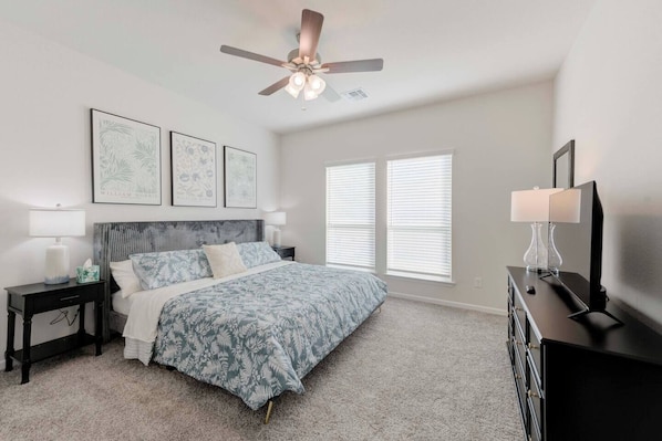 A bedroom with a bed, a nightstand, a dresser, a ceiling fan, and two windows with blinds.