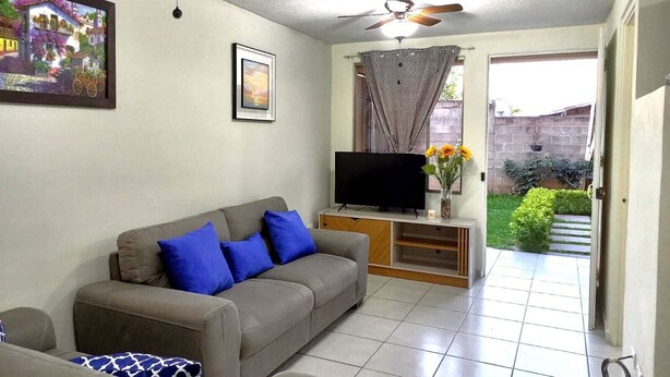 A living room with a grey sofa, blue cushions, a flat-screen TV on a wooden stand, a ceiling fan, and a painting on the wall.