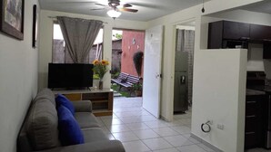 A living room with a sofa, TV, and a ceiling fan. There is a kitchen area with cabinets and a refrigerator.