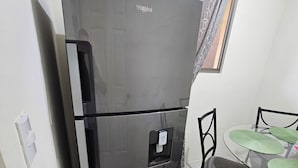 A compact kitchen with a black Whirlpool refrigerator, a dining table with green placemats, and a window with patterned curtains.