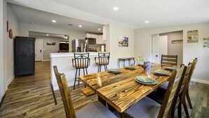 A dining area with a wooden table and chairs, a kitchen in the background, and a dark door on the left. - Diamond in the Ruff -4bdrm Hot Tub / Firepit / Boat Parking Broken Bow Hochatown (Broken Bow)