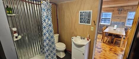 A compact bathroom with a shower, toilet, and sink. A wooden door leads to a dining area with a table and chairs.