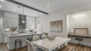 A modern kitchen with white cabinets, a central island, and a dining area with a white table and chairs. - Pura Vida by Avantstay 30A Treetop Retreat Shared Pool + Private Beach Access + Bikes (Panama City Beach)