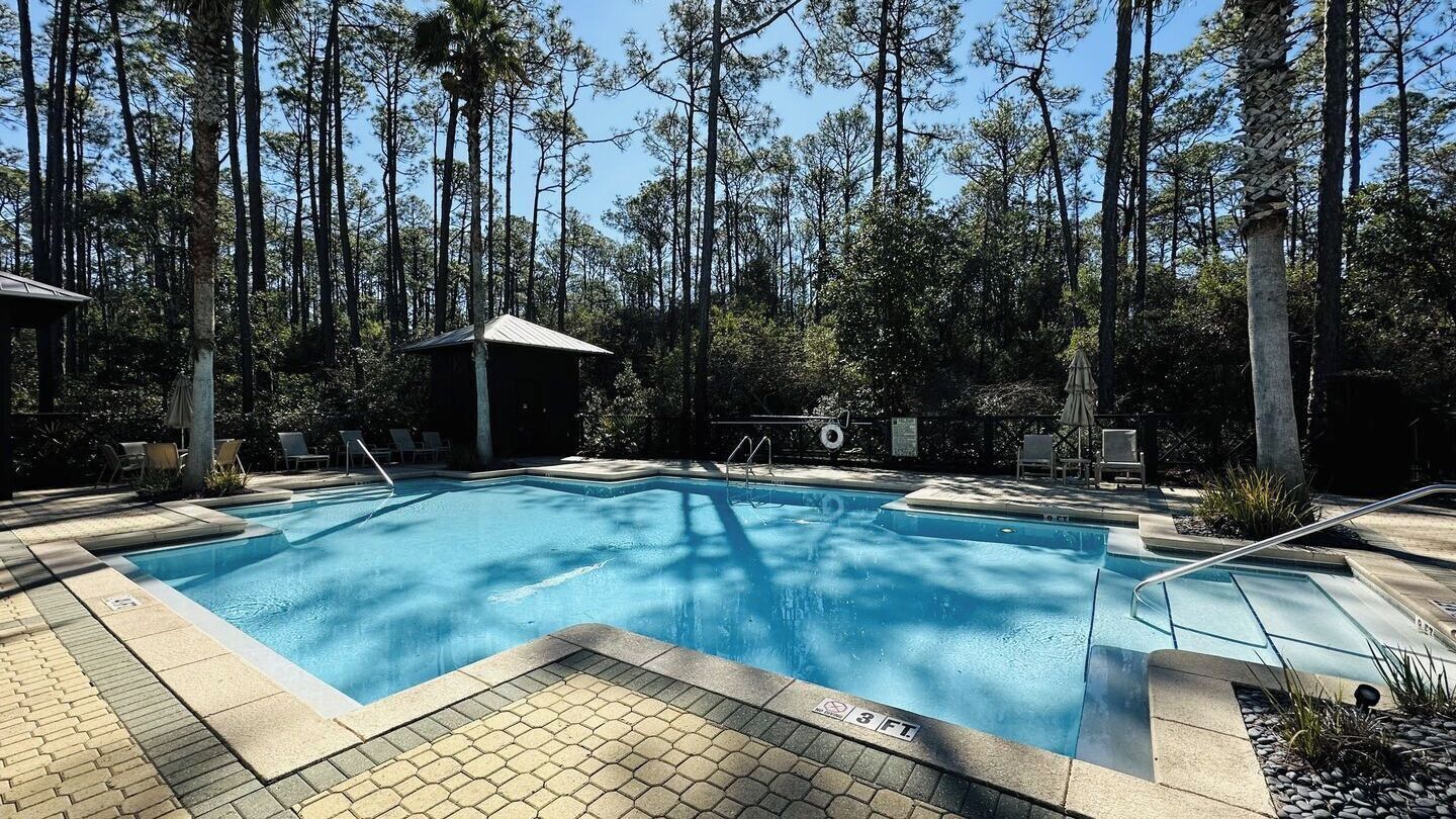 Une piscine rectangulaire, bordée d’une terrasse carrelée, entourée d’arbres et dotée d’un espace salon abrité.
