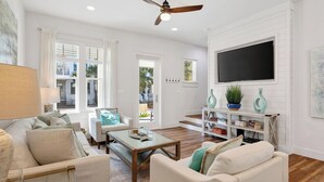A spacious living room with a ceiling fan, a flat-screen TV, and a bookshelf. - Pura Vida by Avantstay 30A Treetop Retreat Shared Pool + Private Beach Access + Bikes (Panama City Beach)