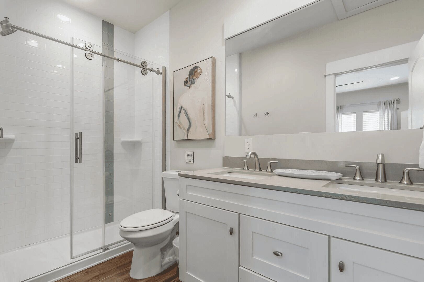 Une salle de bain moderne avec une douche à l’italienne en verre, des toilettes, un lavabo avec un miroir au-dessus et un tableau accroché au mur.