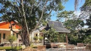 An outdoor seating area with wicker furniture and a fire pit, surrounded by trees and a building with a sloped roof. - Dreamz Village (Langkawi)