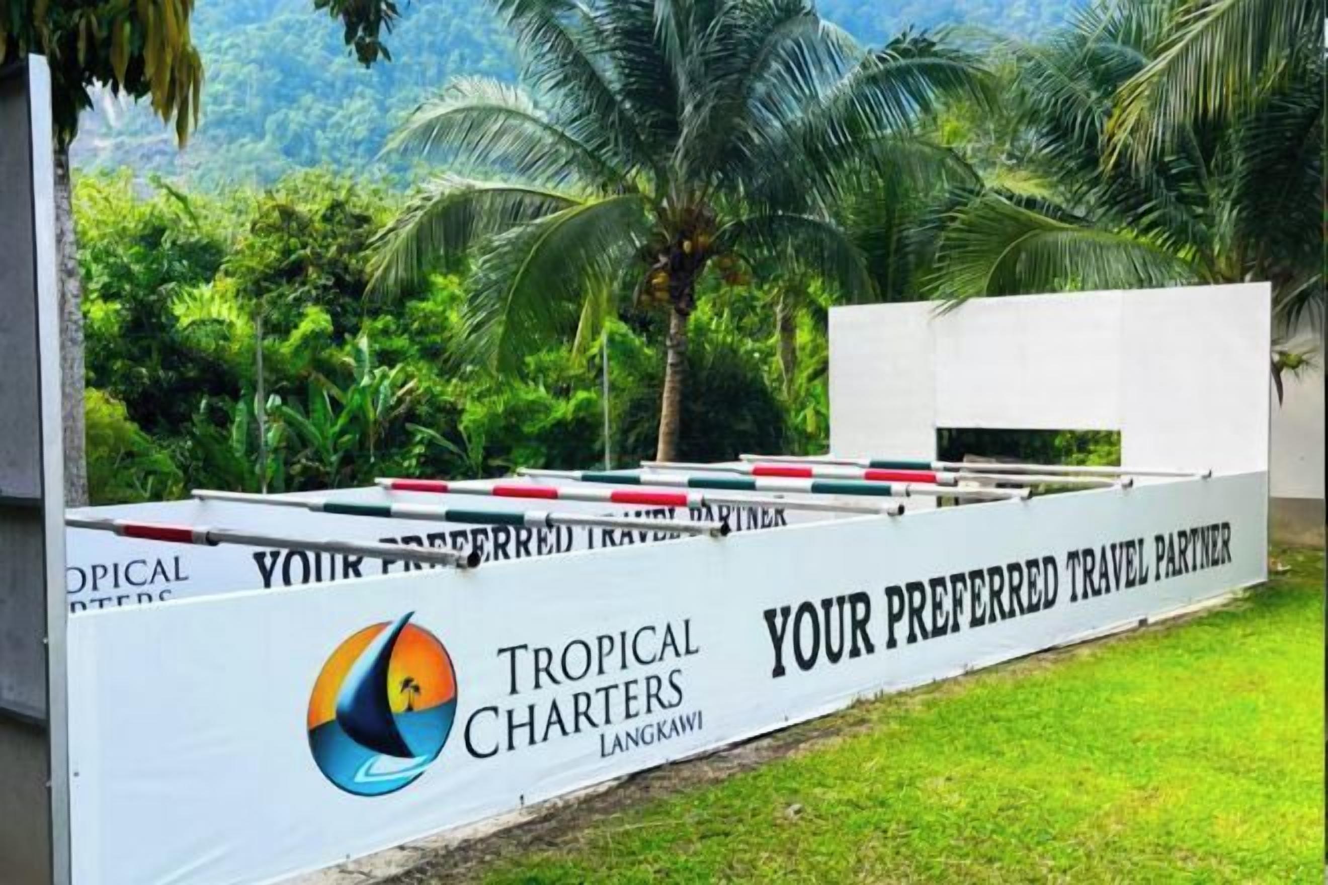A white sign with 'Tropical Charters Langkawi' and 'Your Preferred Travel Partner' text, set against a backdrop of greenery and palm trees.