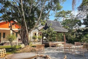 An outdoor seating area with wicker furniture and a fire pit, surrounded by trees and a building with a tiled roof. - Dreamz Village (Langkawi)