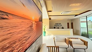A modern hotel lobby with a large abstract painting, a reception desk, and a seating area with a view of the outdoors. - Makia Beach Experience (Ipojuca)